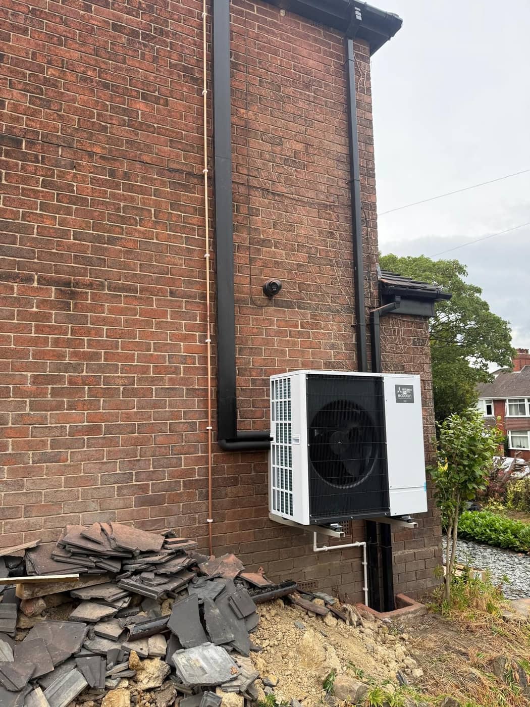 A white air source heat pump beside a tall brick gable wall under a cloudy sky