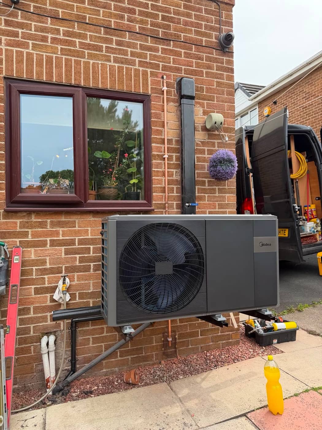An air source heat pump on wall brackets beside a front window, with the engineer's van and tools behind