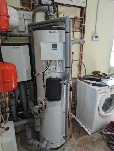 Hot water tank with pipes and a thermostat, next to a washing machine.