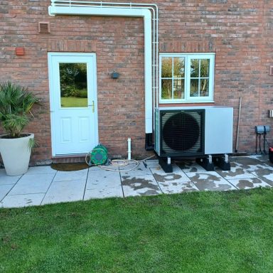 Heat pump installed beside a door, with a small plant and patio area in view.