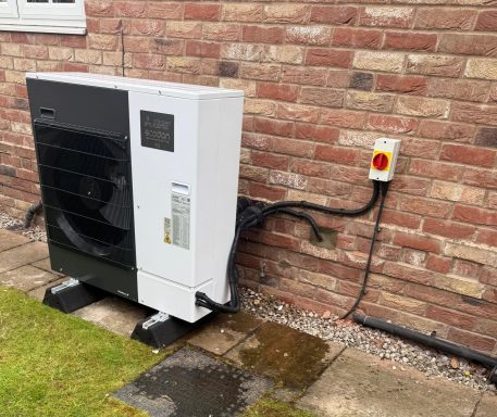 Air source heat pump mounted on a brick wall, with a power disconnect switch nearby.