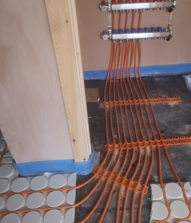 Orange heating pipes arranged on a floor with circular tiles and a wall in the background.