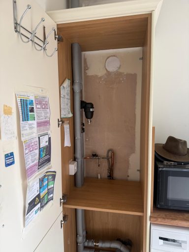 Empty cupboard with exposed pipes and bare wall, alongside a small kitchen area.